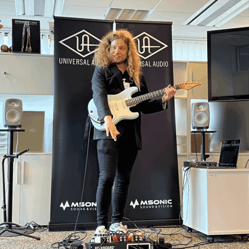 Markus Venehsalo sähkökitara kaulassa ja osoittamassa sormella lattialla olevia Universal Audion UAFX-pedaaleja.