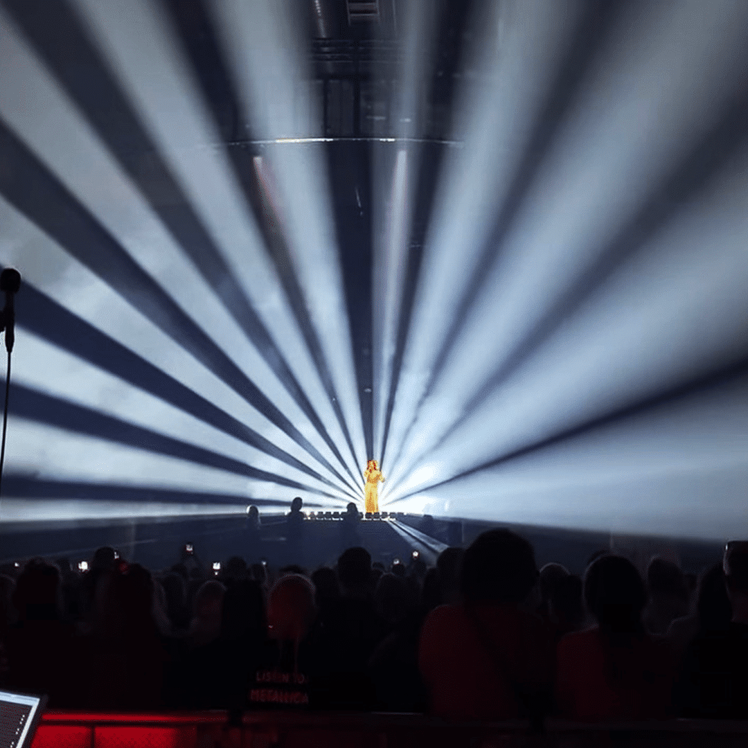 Stiina Jean lavalla laulamassa Nylon Beatin Tytöt haluaa -konsertissa. Hänen takaa loistaa erittäin kirkas valkoinen valo kohti yleisöä ja kattoa.
